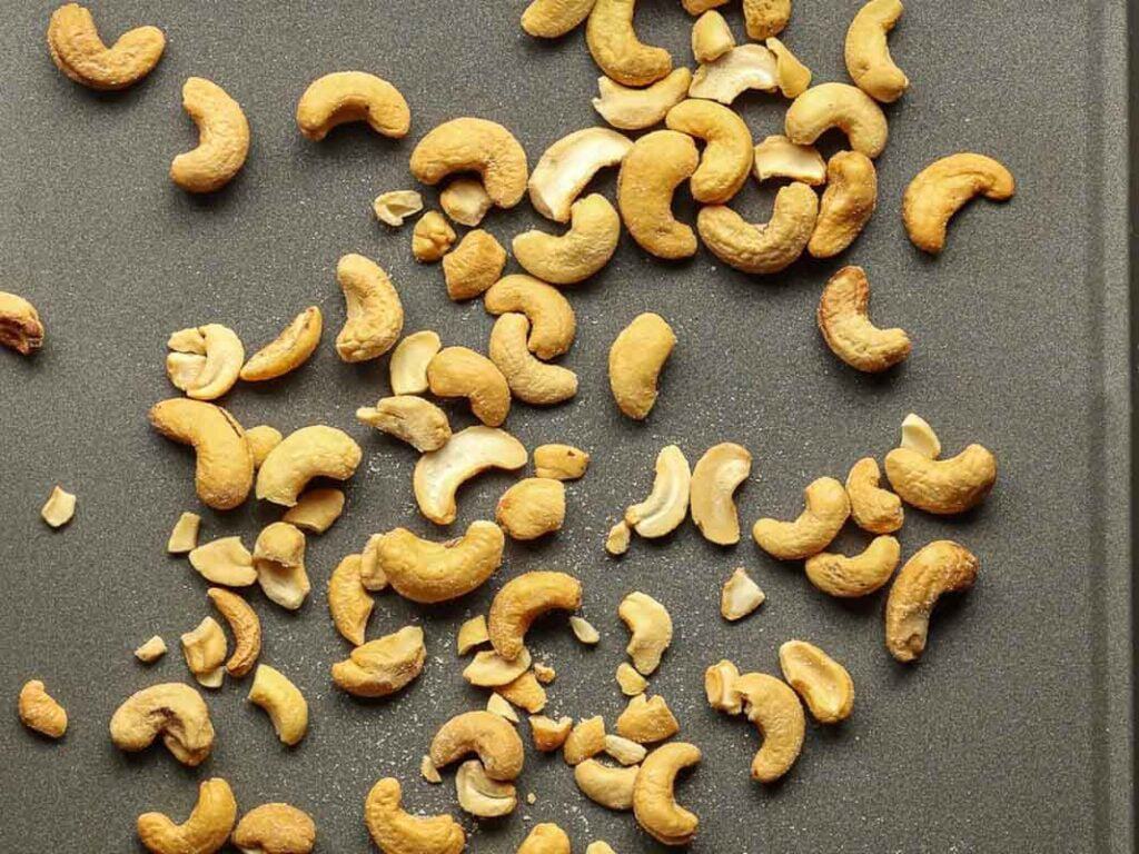 Scattered roasted cashews on a flat grey surface, showcasing various whole nuts and some broken pieces.